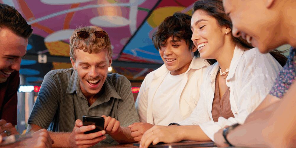 Group of friends laughing and smiling as they look at a phone together in a vibrant, colourful setting.