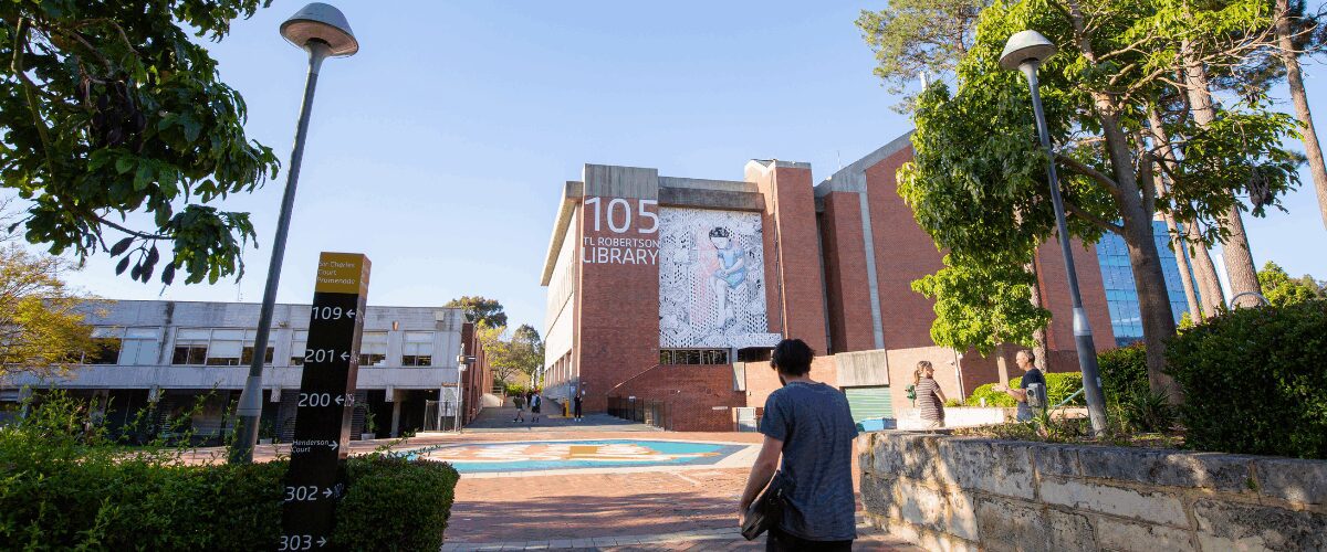Student walking towards the Curtin University Library, with the building number “105” displayed on the wall and trees surrounding the area.