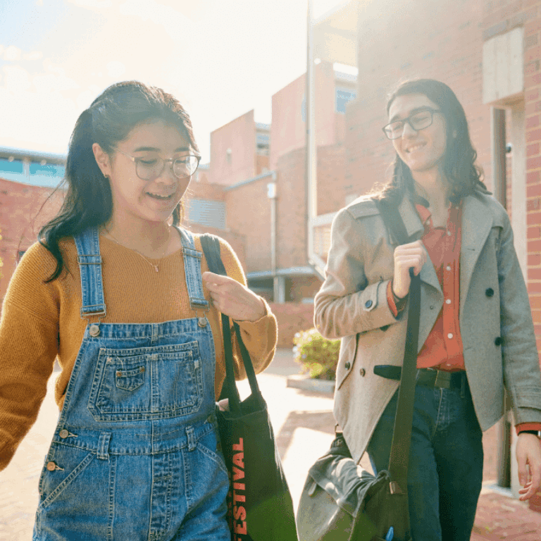 Two Curtin students walking past Building 407, Business and Law, carrying bags and chatting in the sunshine.