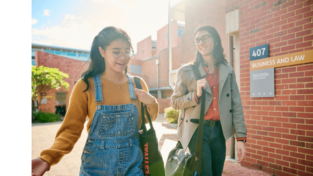 Two Curtin students walking past Building 407, Business and Law, carrying bags and chatting in the sunshine.
