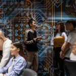 Students working at computers and discussing in a classroom with a large circuit board artwork on the wall.