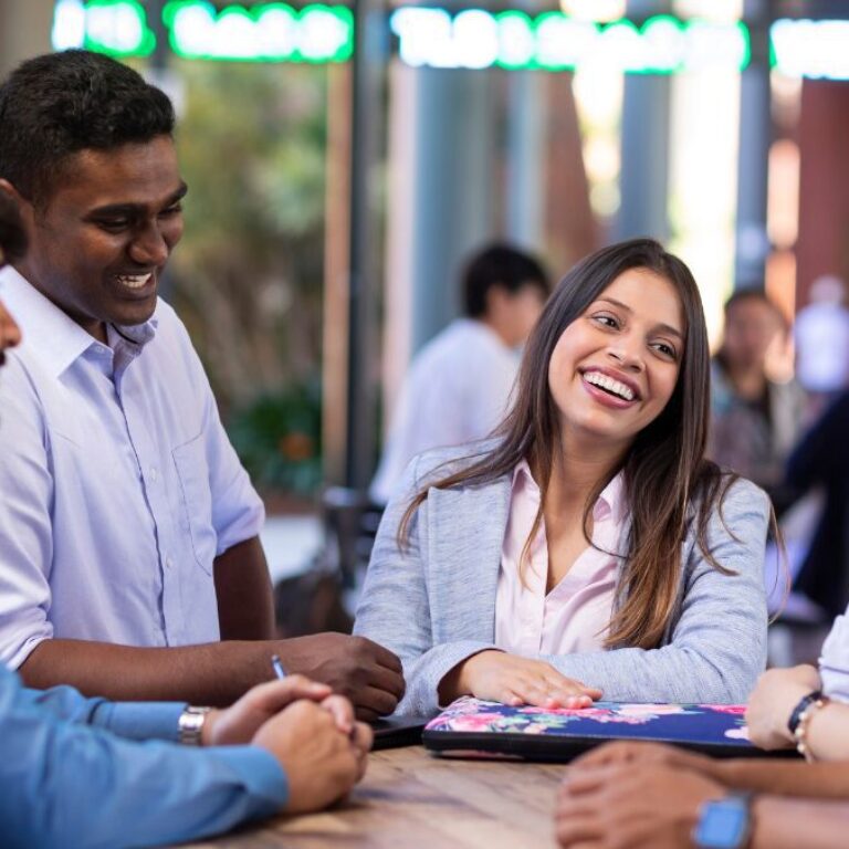 Group of university students gathered around a table, smiling and talking together in an outdoor setting.