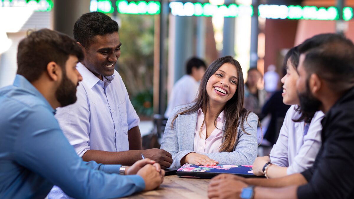 Group of university students gathered around a table, smiling and talking together in an outdoor setting.