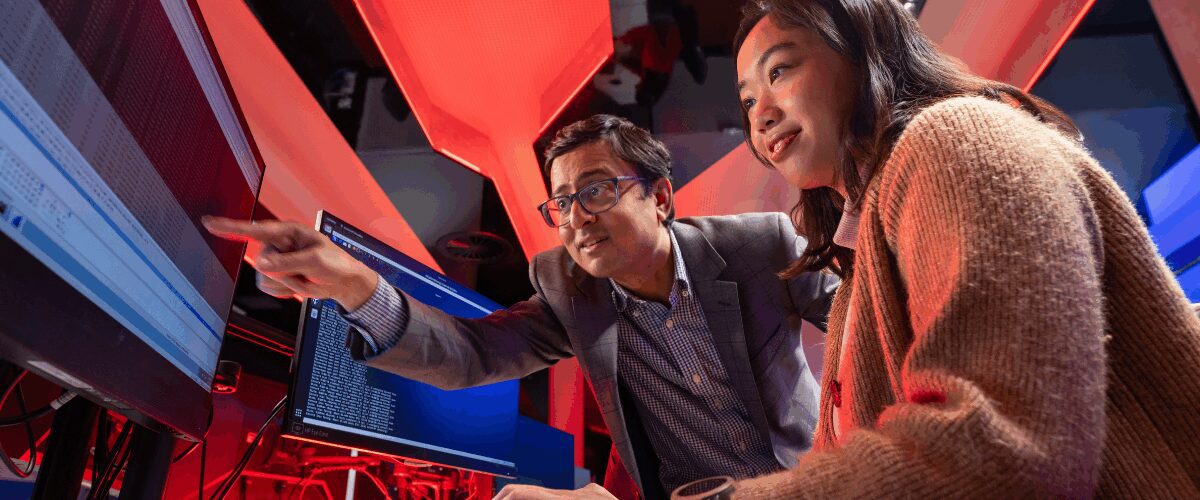 University student working at a computer with a lecturer pointing at the screen in a modern, red-lit lab.