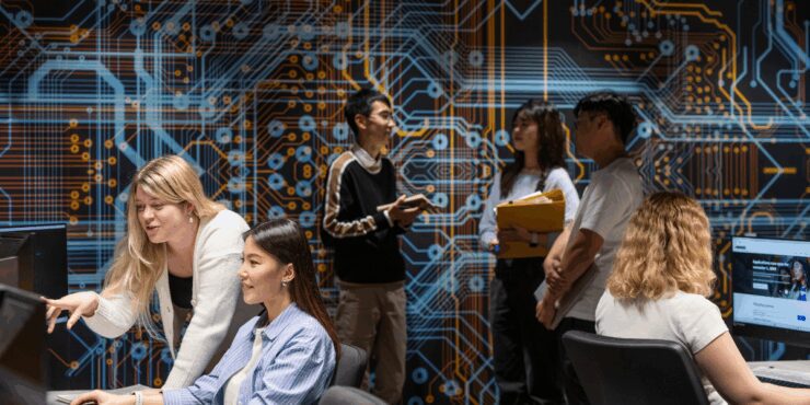 Students working at computers and discussing in a classroom with a large circuit board artwork on the wall.