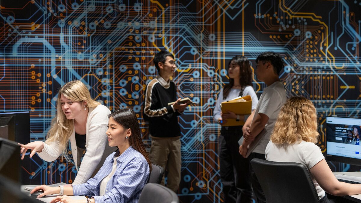 Students working at computers and discussing in a classroom with a large circuit board artwork on the wall.