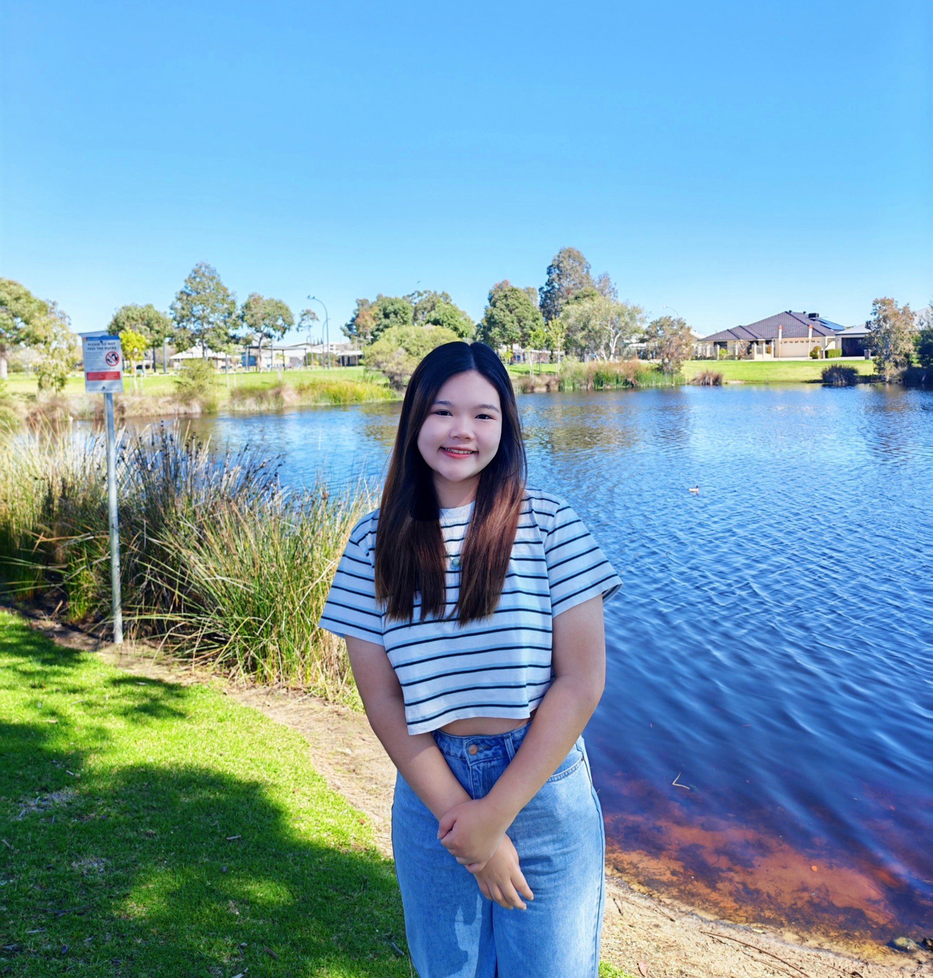 Person standing and smiling outdoors. Behind is a lake and greenery. Above is a bright blue sky.