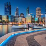 Perth city skyline at dusk, with high-rise buildings lit up and reflections on the water at Elizabeth Quay.