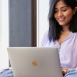 Person using a silver laptop on a red chair near a window.