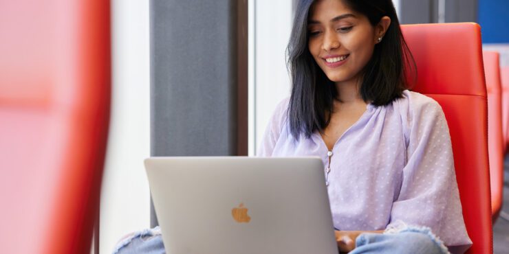 Person using a silver laptop on a red chair near a window.