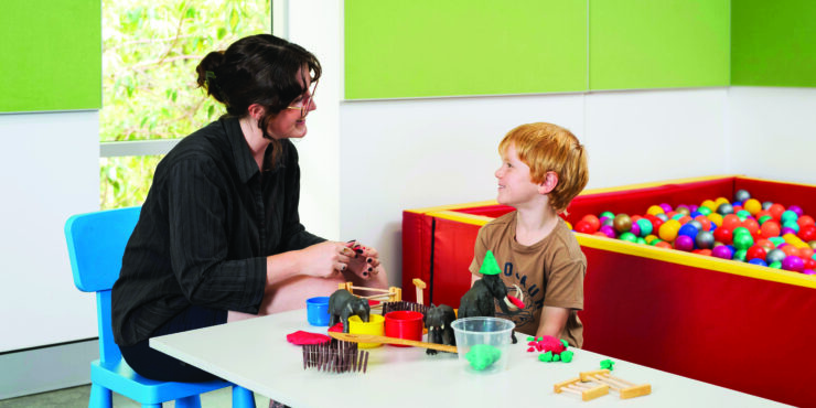 A woman teacher sits at a tabe with a young boy with a game in front of them and a ball bit behind them.