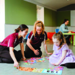 Two female student occupational therapists sit on the floor with a young child playing a card game.