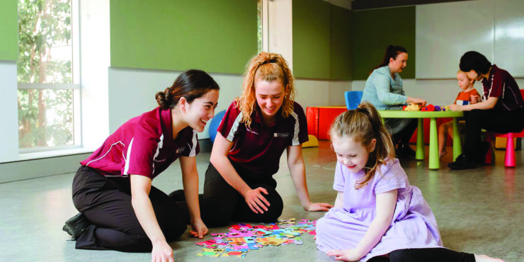 Two female student occupational therapists sit on the floor with a young child playing a card game.