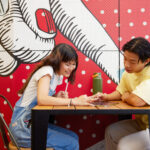 Two people sitting at a table looking at something together, with a colourful mural featuring hand illustrations behind them.