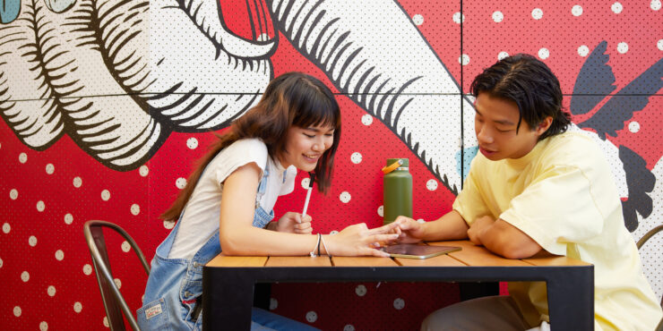 Two people sitting at a table looking at something together, with a colourful mural featuring hand illustrations behind them.