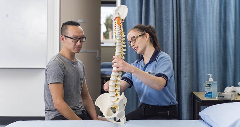 An OT student points to a replica of a spine while speaking to a patient.
