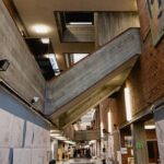 Interior of a modern building with exposed concrete and brick walls, angled staircases, and architectural drawings displayed along the corridor.