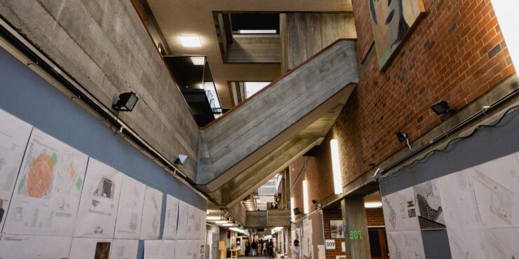 Interior of a modern building with exposed concrete and brick walls, angled staircases, and architectural drawings displayed along the corridor.