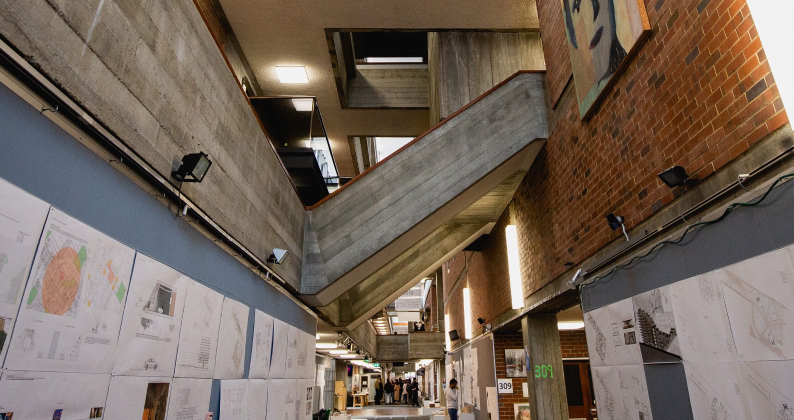 Interior of a modern building with exposed concrete and brick walls, angled staircases, and architectural drawings displayed along the corridor.