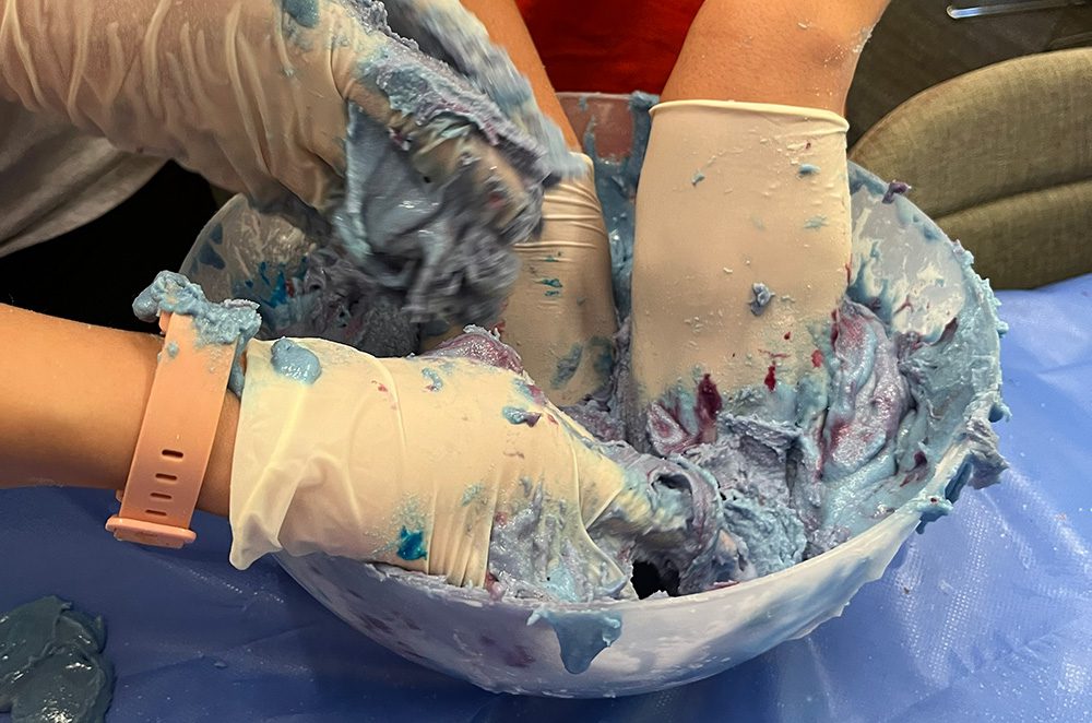 Gloved hands mixing blue matter in a bowl.