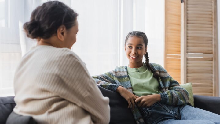 Two people sitting on a couch engaged in conversation, one smiling at the other.