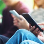 Three people sitting closely together, each holding and using a smartphone, with a blurred background
