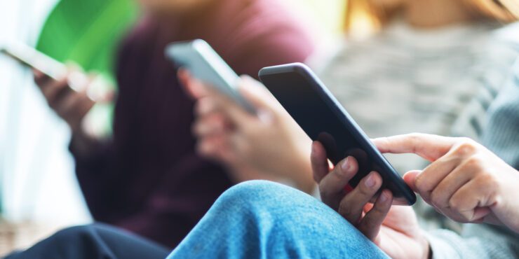 Three people sitting closely together, each holding and using a smartphone, with a blurred background