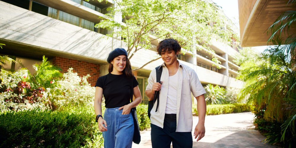 Two people walking outdoors along a paved path surrounded by greenery and modern buildings, one wearing a black top and blue jeans, the other carrying a backpack.
