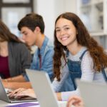 Portrait of smiling university student sitting in a row with classmates, who are all studying together on laptops.