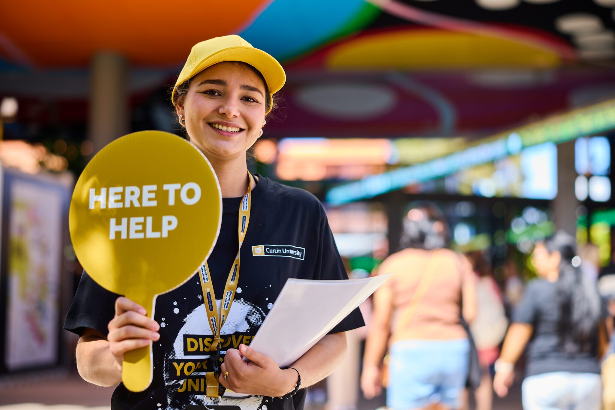 A staff member wearing a yellow cap holds a yellow ‘Here to Help’ sign while standing at an outdoor campus event, carrying papers and surrounded by colourful decorations and people walking by.