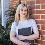 Student holding a laptop standing in front of a brick wall