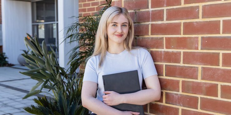 Student holding a laptop standing in front of a brick wall