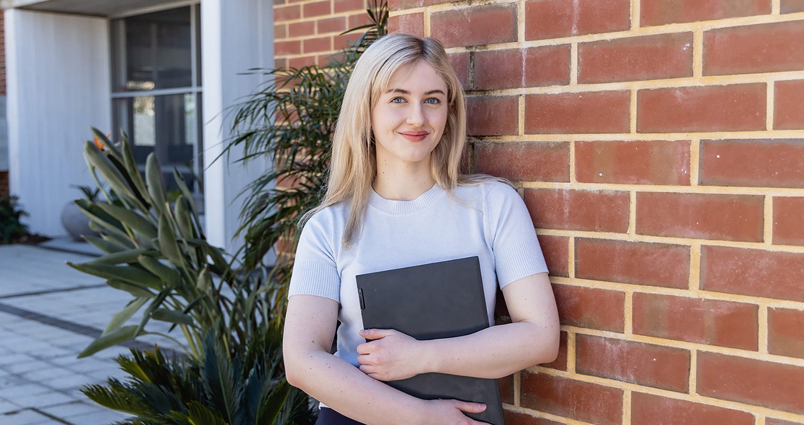 Student holding a laptop standing in front of a brick wall