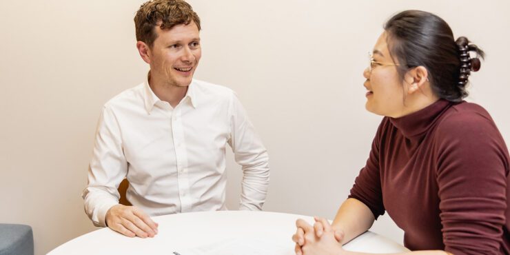 Two people seated at a small table in an office, having a face‑to‑face conversation with papers and a pen between them.