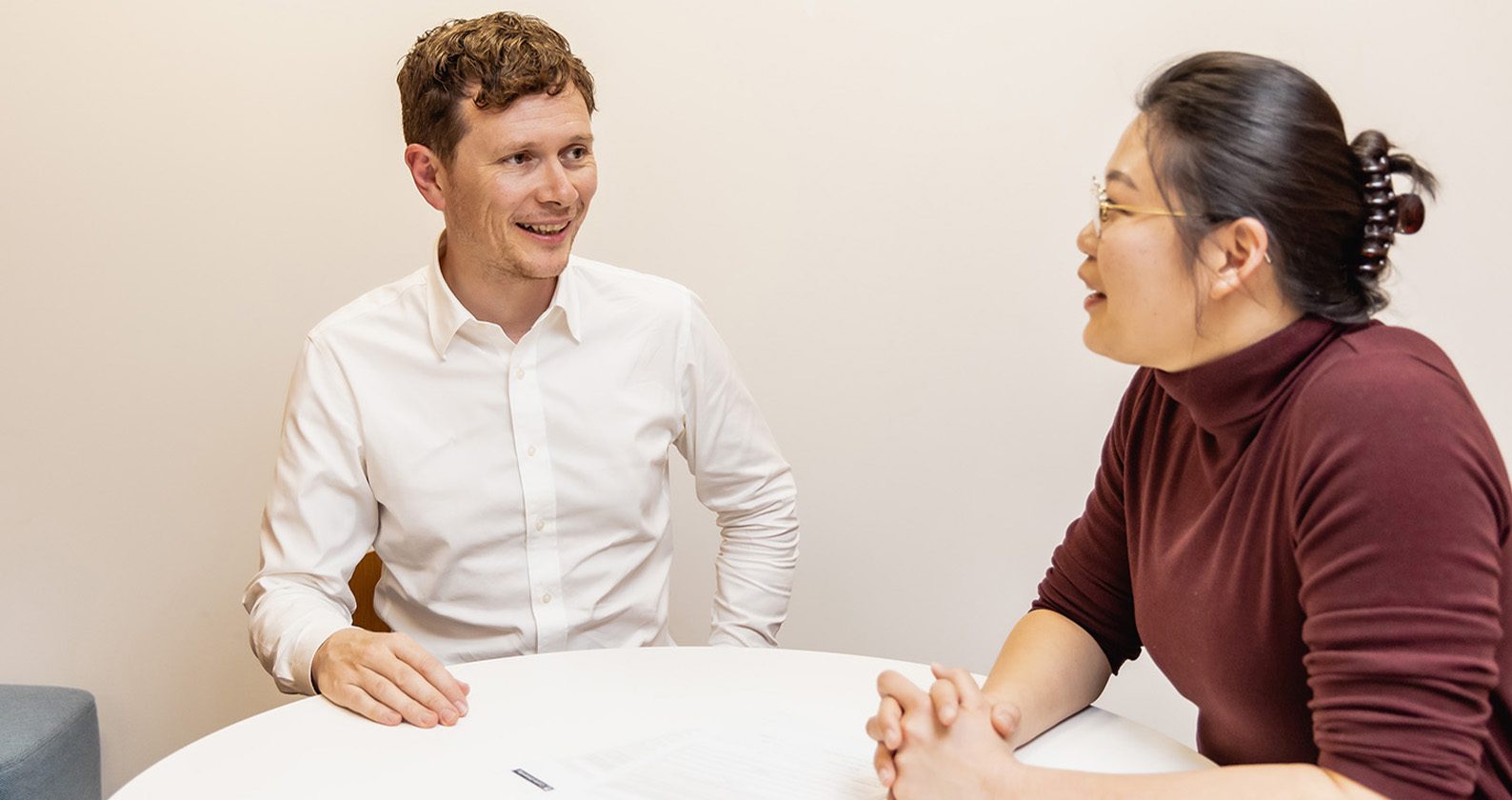 Two people seated at a small table in an office, having a face‑to‑face conversation with papers and a pen between them.