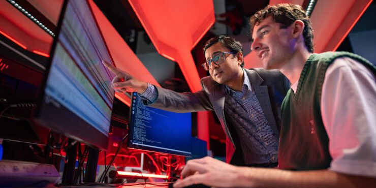A young man sits at computer with teacher standing next to him and pointing at the computer screen.