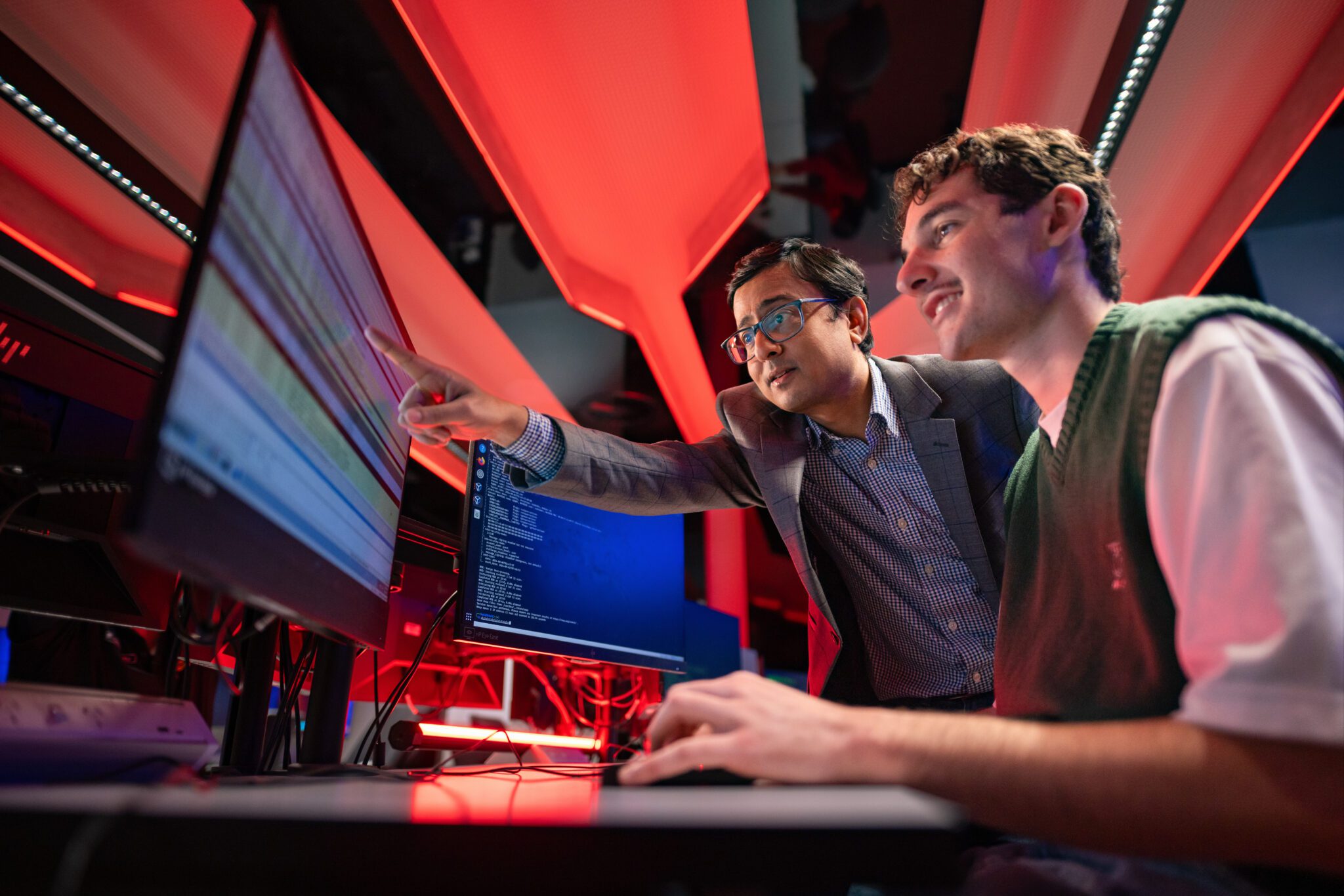 A young man sits at computer with teacher standing next to him and pointing at the computer screen.