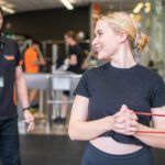 Student performing a resistance band exercise in a university gym with a trainer nearby.