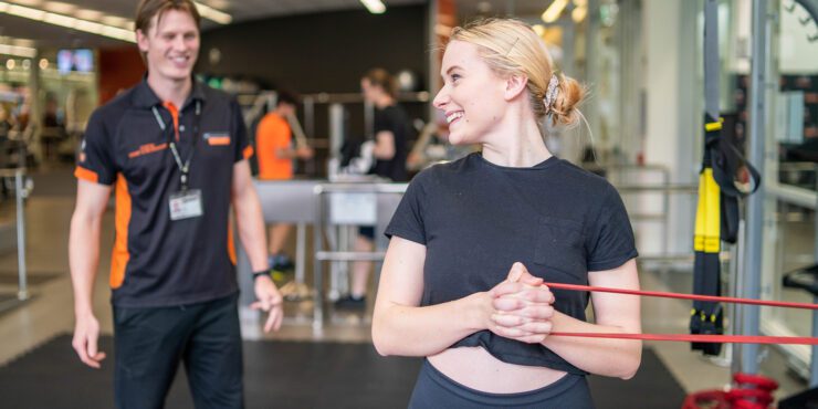 Student performing a resistance band exercise in a university gym with a trainer nearby.