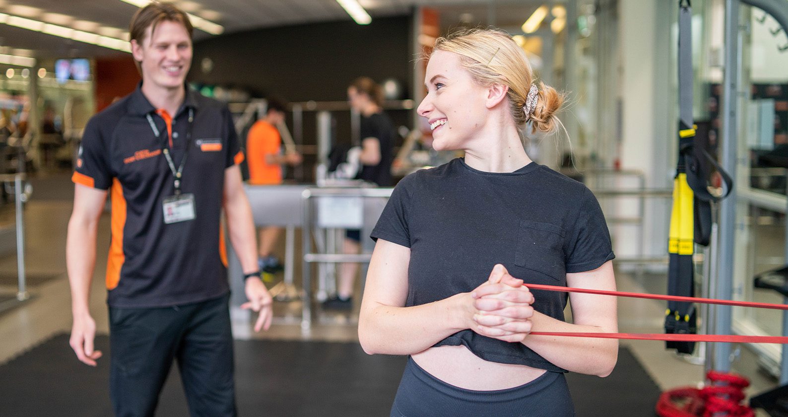 Student performing a resistance band exercise in a university gym with a trainer nearby.