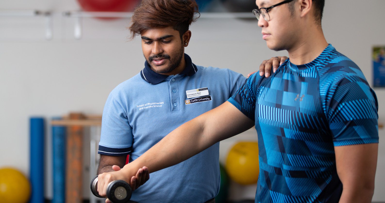 Physiotherapy student guiding a client through an arm strength exercise with a dumbbell in a clinic setting.