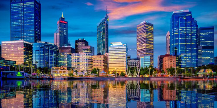 City skyline at dusk with illuminated high‑rise buildings reflected on calm water under a colourful evening sky.