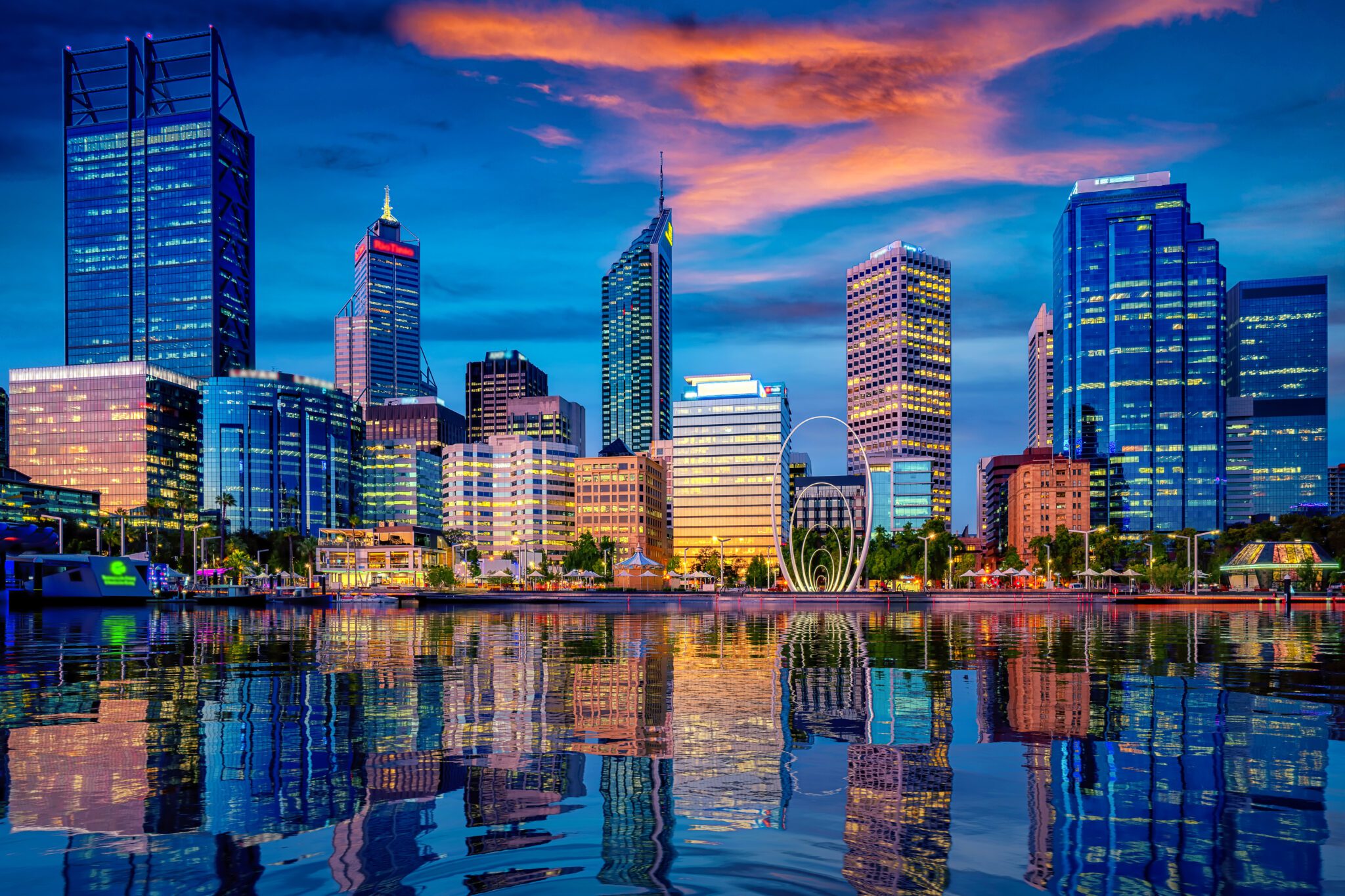 City skyline at dusk with illuminated high‑rise buildings reflected on calm water under a colourful evening sky.
