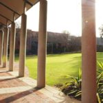 Covered walkway with columns beside a grassy campus courtyard in sunlight.
