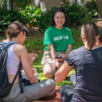 Students sitting on grass in a small group, chatting with a mentor wearing a “Mentor” T‑shirt.