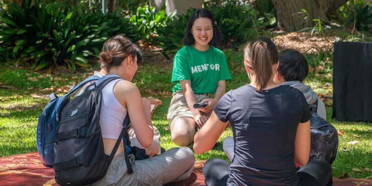 Students sitting on grass in a small group, chatting with a mentor wearing a “Mentor” T‑shirt.