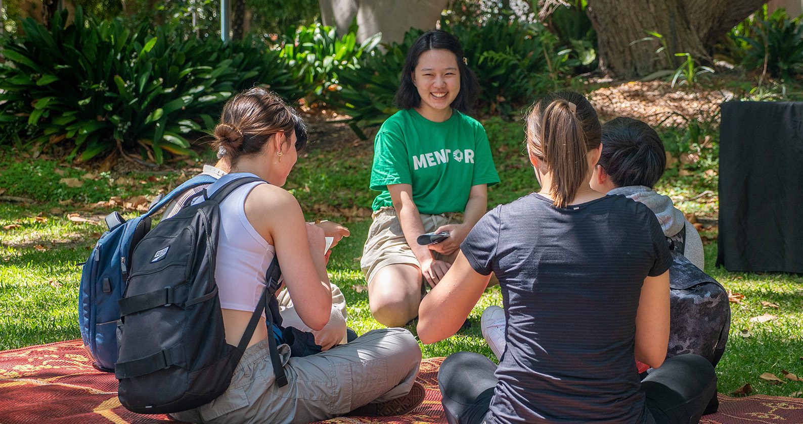 Students sitting on grass in a small group, chatting with a mentor wearing a “Mentor” T‑shirt.