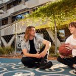 Two people sitting on an outdoor basketball court holding a basketball, with campus buildings in the background.