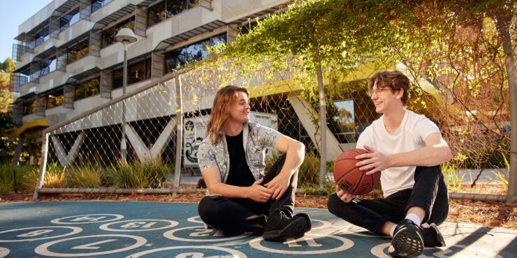 Two people sitting on an outdoor basketball court holding a basketball, with campus buildings in the background.