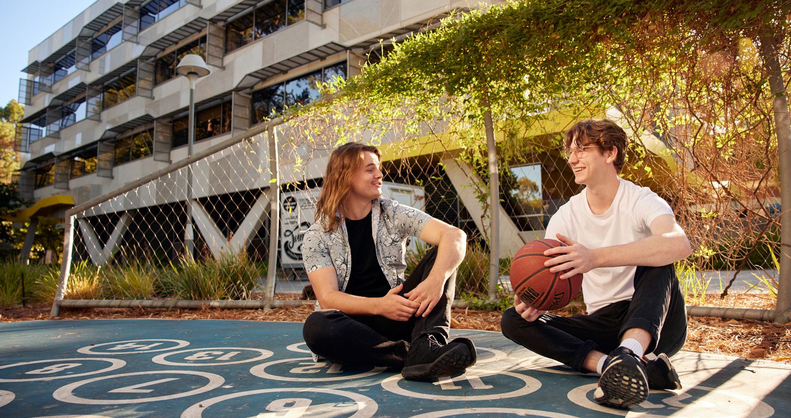 Two people sitting on an outdoor basketball court holding a basketball, with campus buildings in the background.